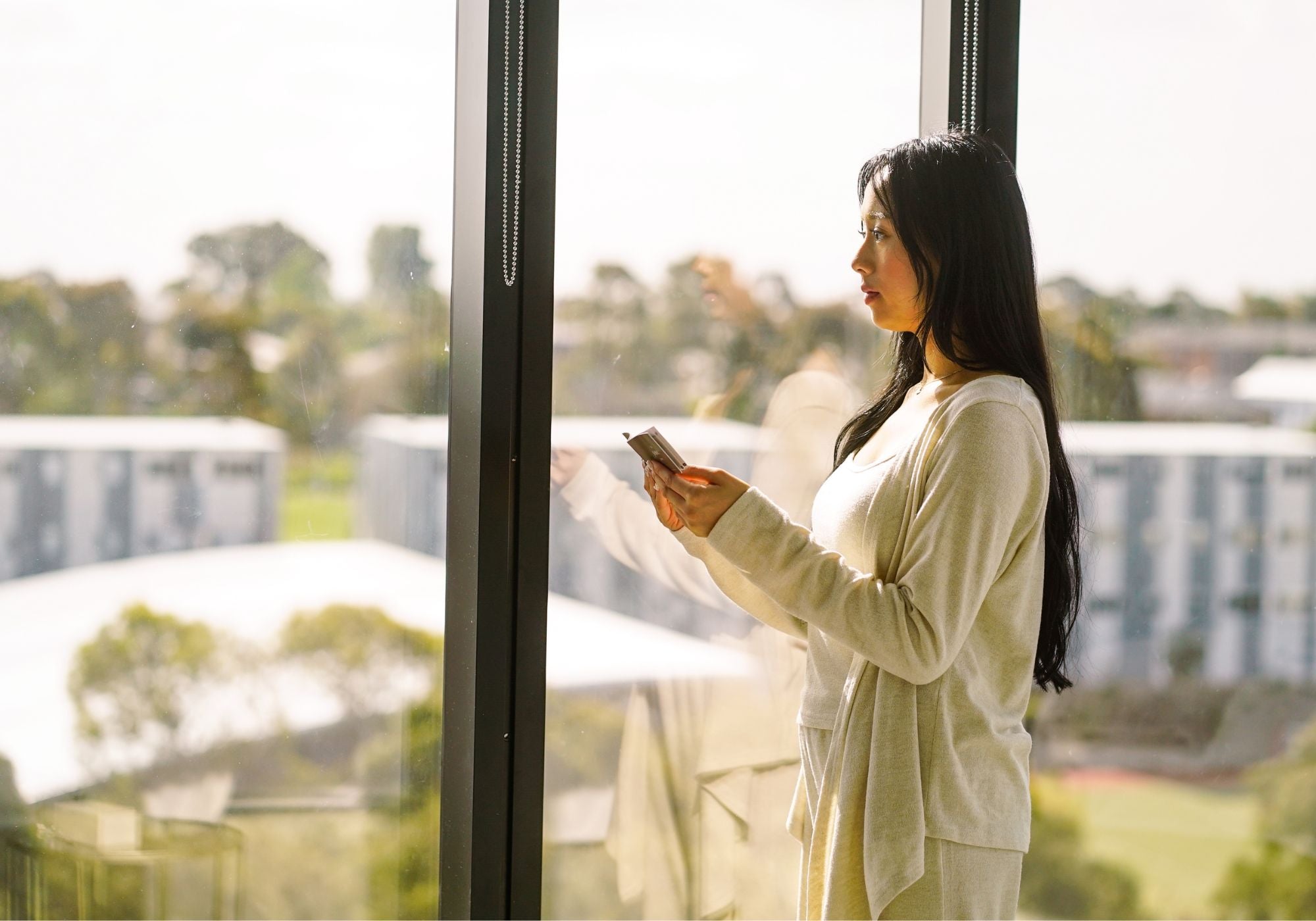 Woman standing by a window reading the Sayang Journey booklet in the morning, showing the softer morning ritual that follows using Sayang Sleep Strips.