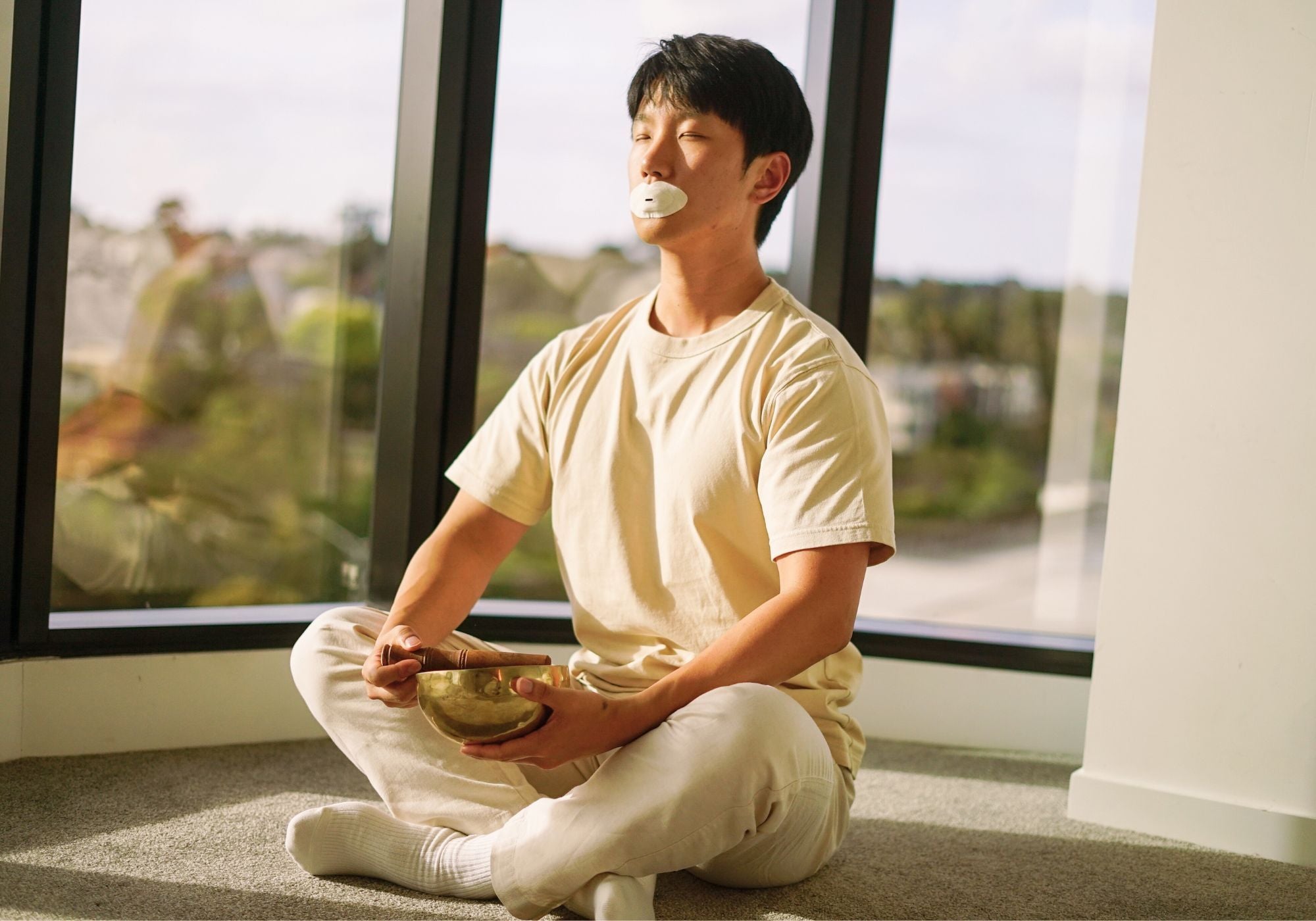 Man sitting cross-legged by a window wearing a Sayang Sleep Strip and holding a meditation bowl during a calm morning ritual.